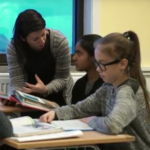 En lærer står ved pulten til en elev. Hun holder en bok i hånda mens hun forklarer noe.