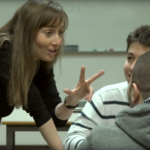 En lærer forklarer elever i klasserommet. Hun holder opp tre fingre.