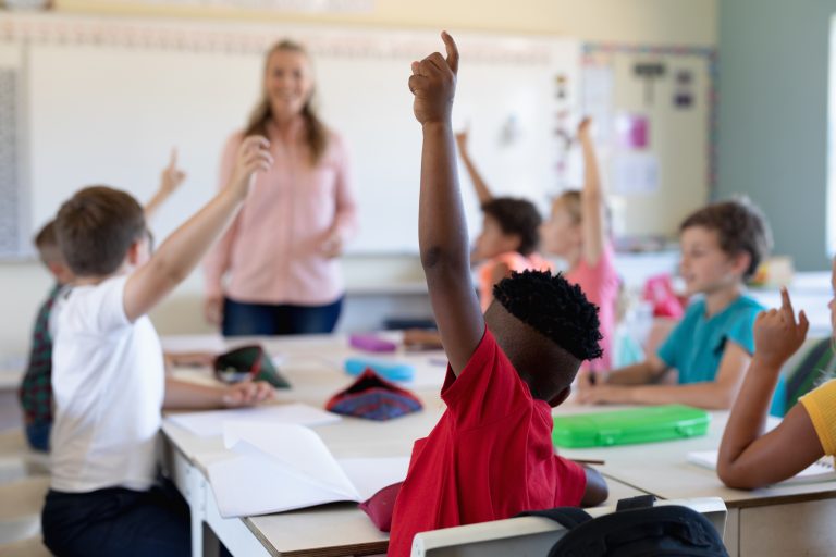 Elever rekker opp hendene i et klasserom. Læreren står ved tavla.
