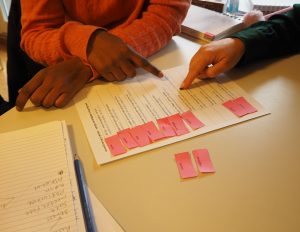 Foto av et ark med setninger. Til venstre for setningene ligger rosa papirlapper med ord på. Hendene til to deltakere er synlig i bildet. De arbeider med arket. 