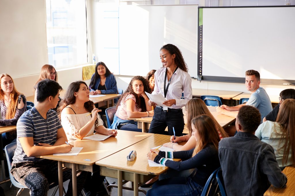 Kvinnelig lærer står midt i et klasserom. Elevene sitter gruppevis.