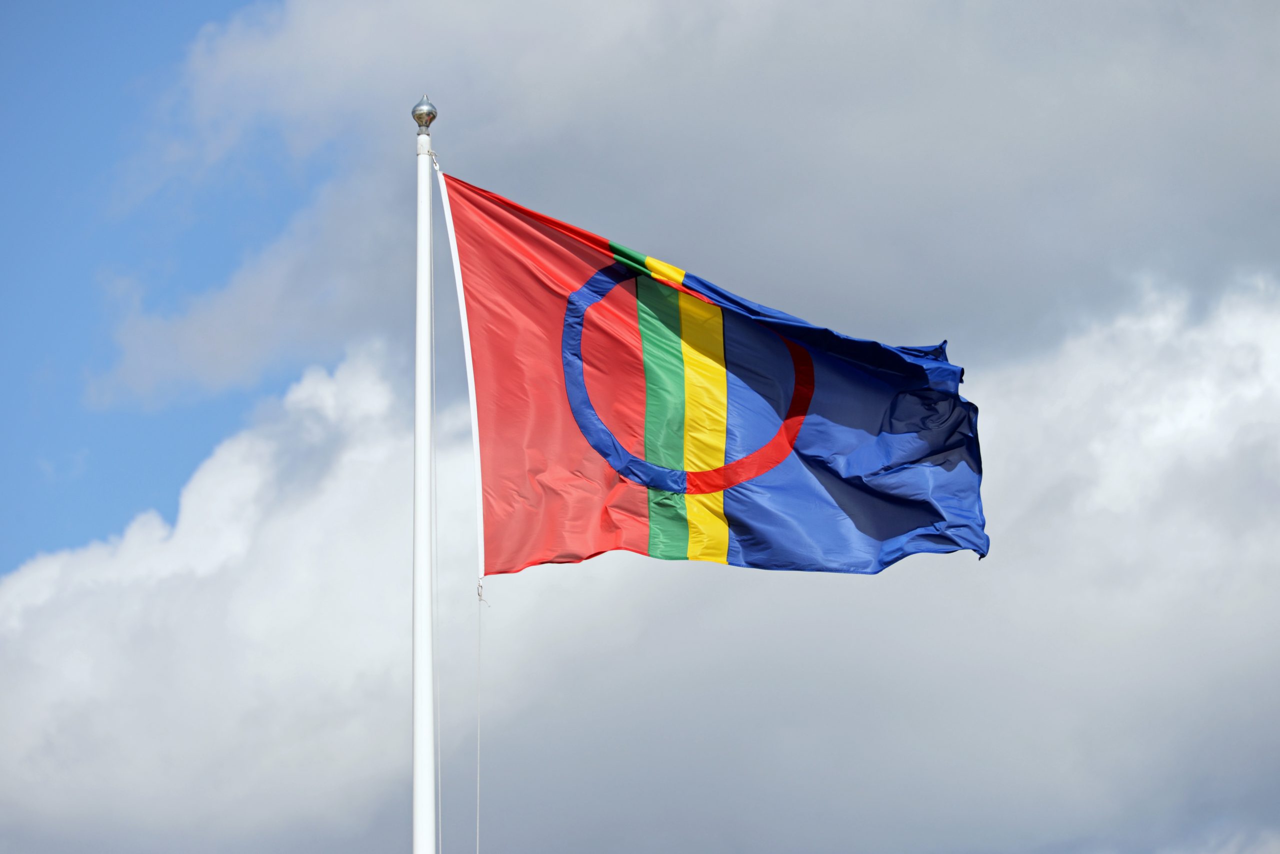 Foto av et samisk flagg som vaier i vinden. På himmelen bak flagget er det hvite skyer.