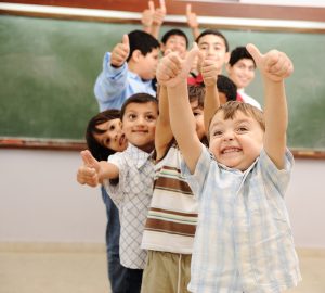Fotografi av skolebarn som står på rekker tomlene opp foran en tavle i et klasserom.