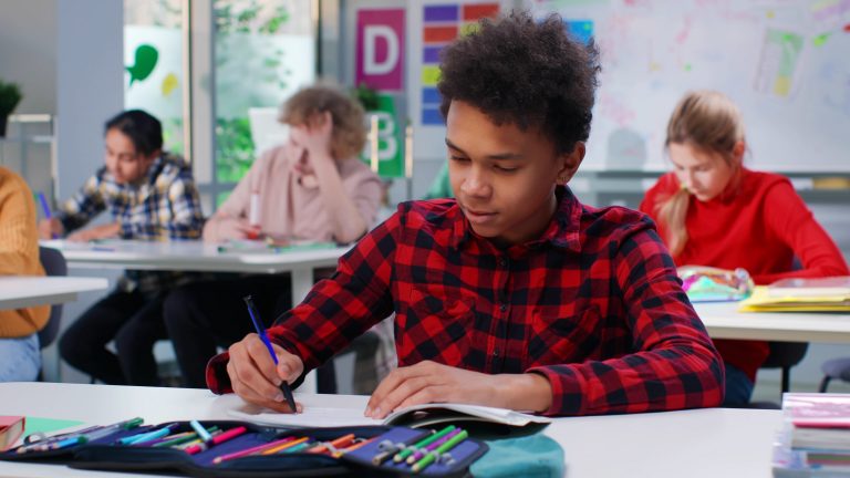 African-American teen student write in notebook at lesson in classroom. Schoolboy study sitting at desk in school
