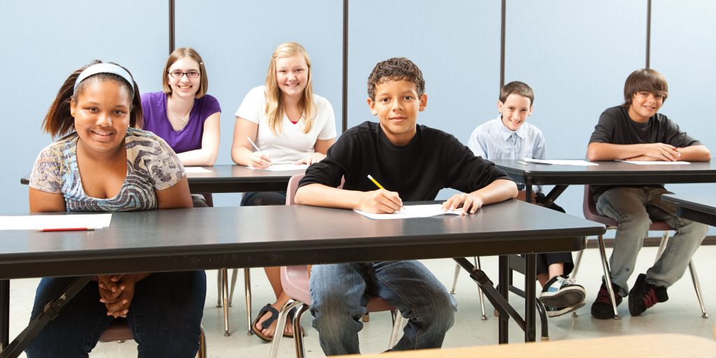 Foto av en innføringsklasse. 6 elever sitter bak bord i et klasserom. Alle ser mot kameraet. De kommer fra ulike steder i verden og har nylig kommet til Norge.