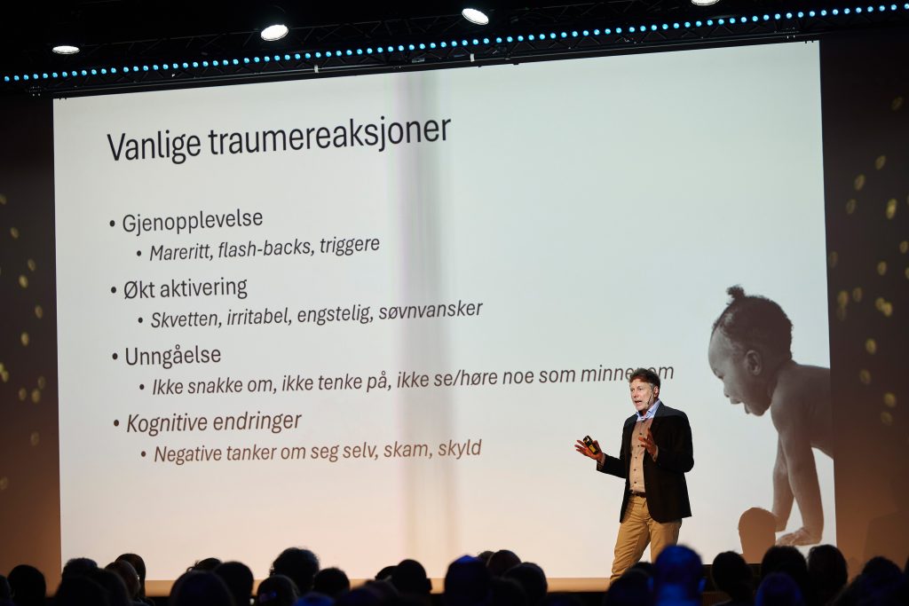 Foto av en person som holder et innlegg på en konferanse. På skjermen bak perosnen står det skrevet: Varige traumereasksjoner