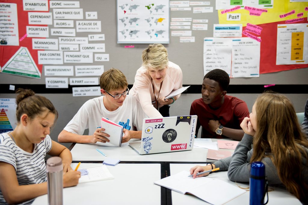 Foto av en gruppe elever i et klasserom. Elevene sitter rundt et bord og jobber. Noen skriver i skrivebøker, mens to elever ser på en PC-skjerm. Bak de to elevene og skjermen står en lærer. Hun peker på skjermen. På veggen bak dem er en stor oppslagstavle. Oppslagstavla er full av plakater, ord, kart og mere.