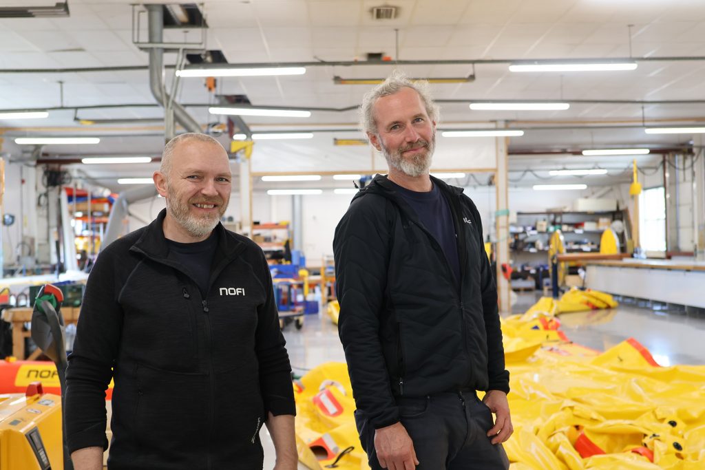 Foto av Geir Olaf Hansen og Anders Larsson. De er i et produksjonsrom. Bak dem ligger en stor gul duk. Det er en oppsamlingsduk for oljesøl som er under produksjon.