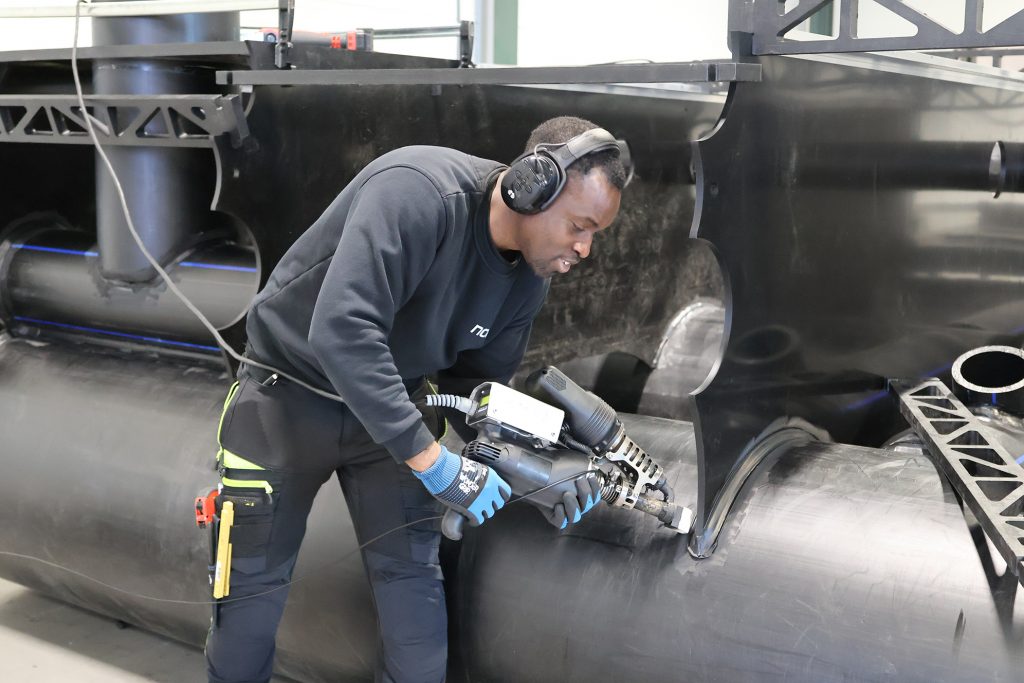 Foto av Tulza Olukan som arbeider på verkstedet. Han har hørselvern på ørene og bruker et elektrisk verktøyg til å utføre en jobb på et stort rør.