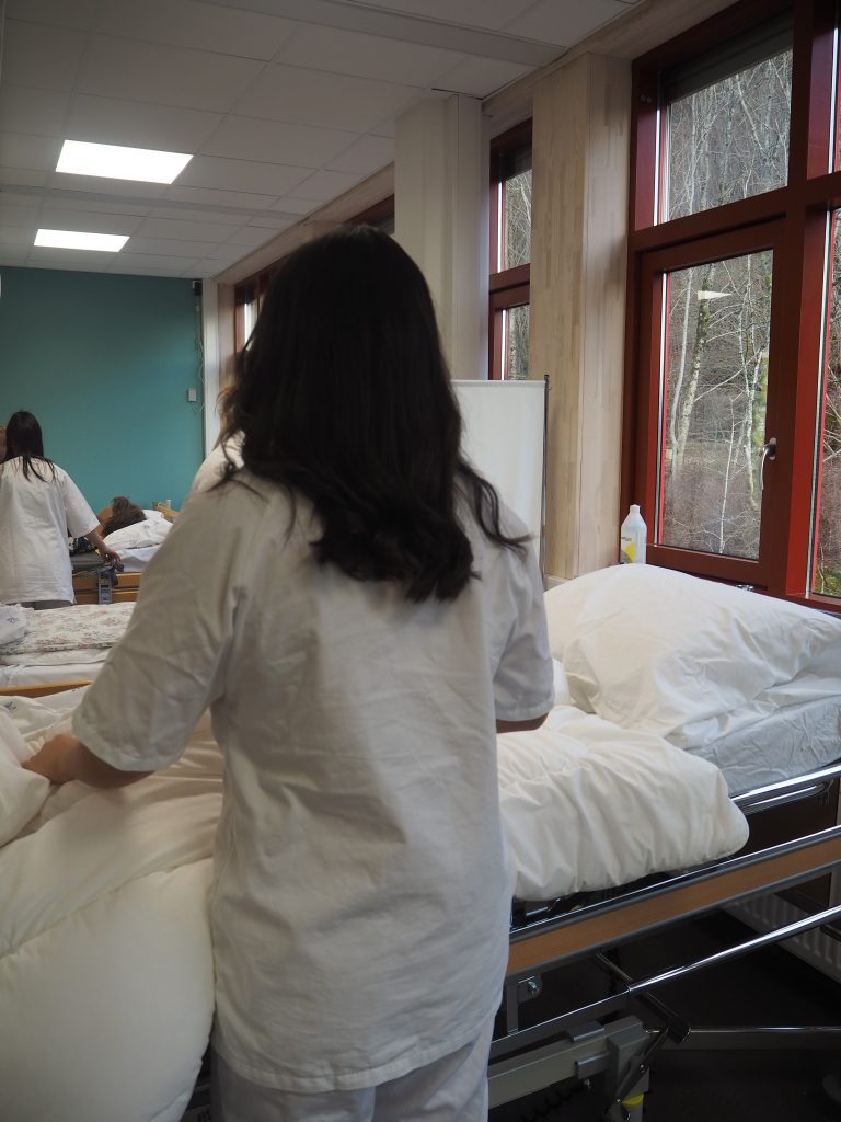 Foto av elever på et øvingsrom på skolen som rer opp en pasientseng. Eleven har hvit uniform på seg og er elev ved helsearbeiderfaget.