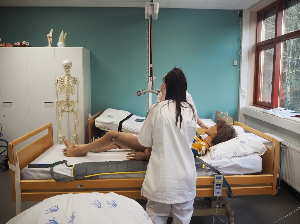 Foto fra et helsefaglig praksis eller øvingsrom på en skole. En elev står ved en seng. I senga ligger en dukke i fullskala størrelse av en voksen. Eleven øver på å bruke en takheis for å forflytte pasienter.