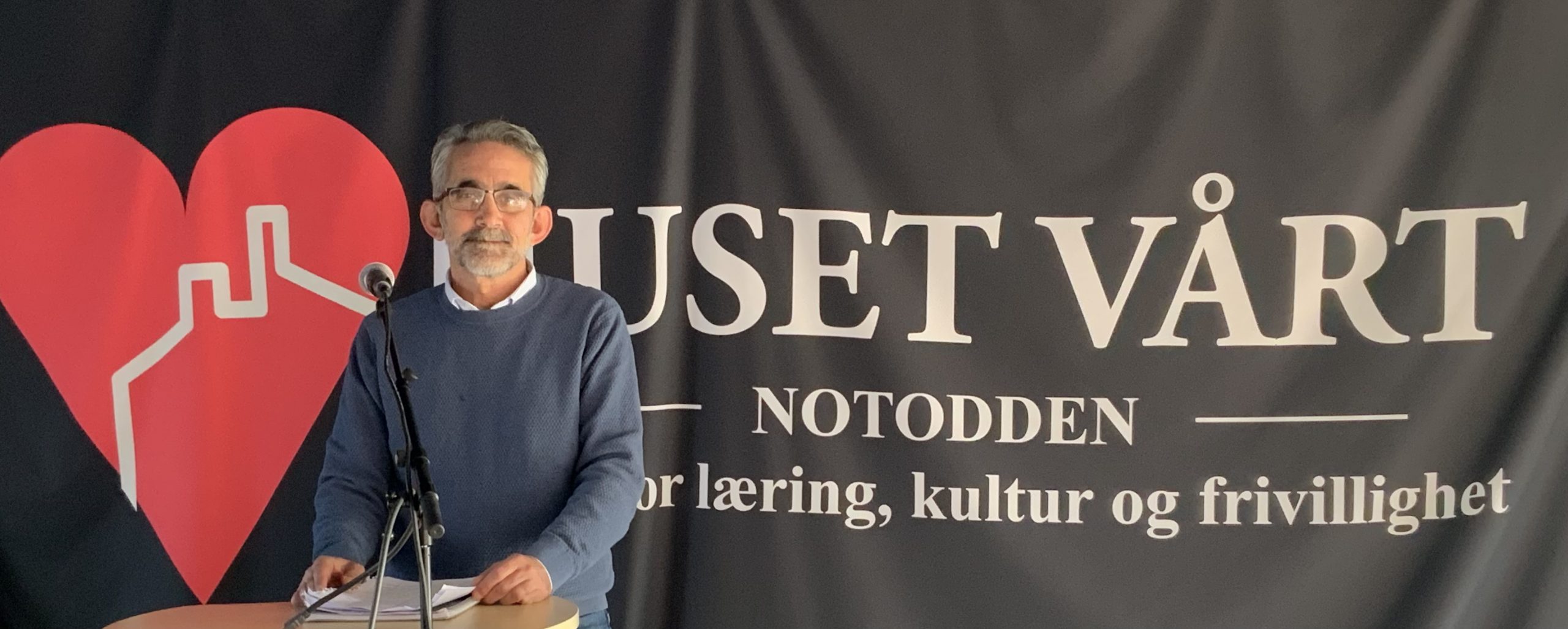 Fotografi av Ali Albakar som står på en talerstol. Bak ham henger det et banner med et stort rødt hjerte og skriften "Huset vårt".