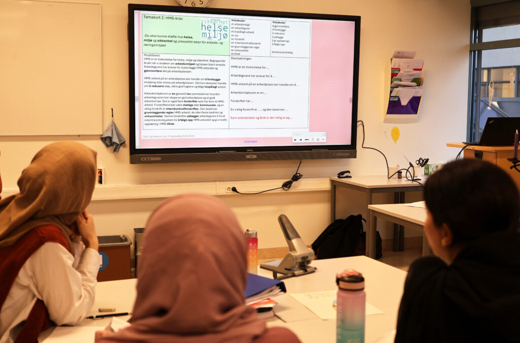 Foto fra et klasserom. En digital tavle er sentral i bildet. På tavla er et temakort om helse, miljø og sikkerhet opp. Tre elever eller deltakere ser mot skjermen