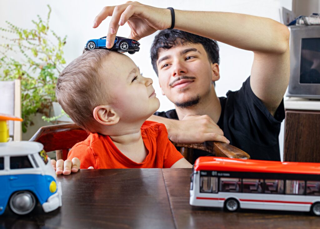 Foto av et barn og en voksen som leker. Den voksne holder en lekebil oppå hodet til barnet. På bordet foran dem stør det to lekebusser.