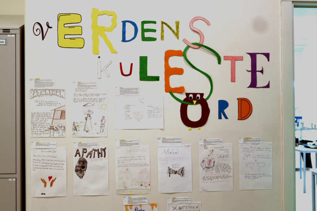 En vegg er dekket med ark med tekst og illustrasjoner. Hvert av arkene inneholder et ord, en forklaring på ordet og en illustrasjon. Over arkene henger en overskrift laget av bokstaver klippet ut i papir. Det står: Verdens kuleste ord.