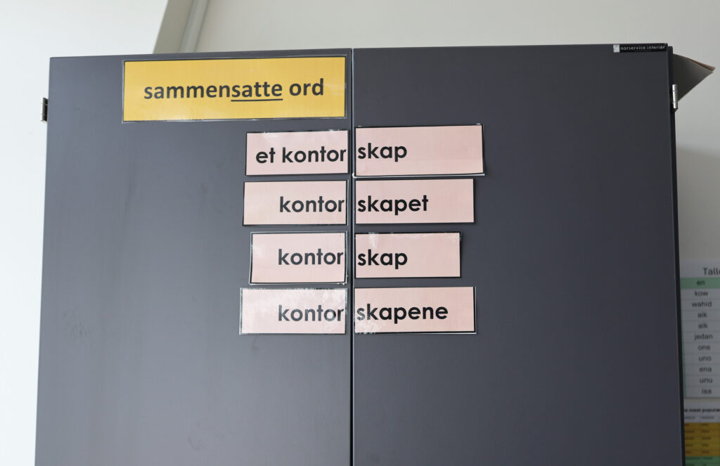Foto av et skap i klasserommet. Øverst på skapet er det skrevet: sammensatte ord. Under, på de to skapdørene er det klistret opp sammensatte ord. På venstred ør står det et kontor, mens det på høyredør står skap. Slik kan en se at kontorskap består av to ord. Videre står det kontorskapet, kontorskap og kontorsakpene.