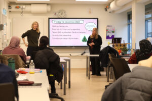 Foto fra et klasserom i voksenopplæringen. Lærerne Hanne Live og Gøril står ved en smart board og underviser en klasse. Noen deltakere sitter ved arbeidsbord og ser mot lærerne. Foto: Katarina Hundal