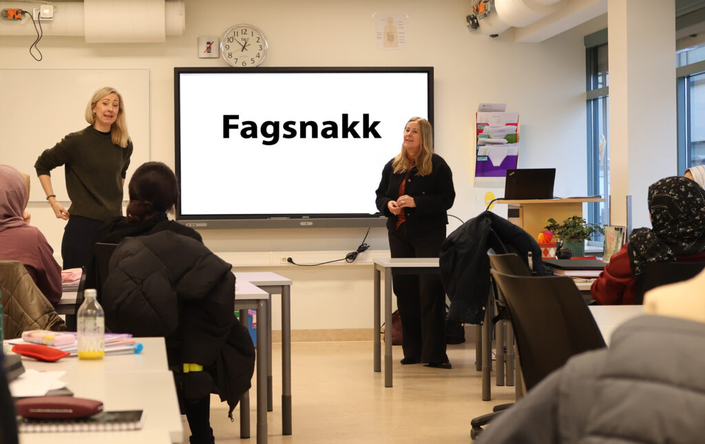 Foto av to lærere som står ved en tavle i et klasserom. På tavla står det fagsnakk. Noen elever ser mot tavla.